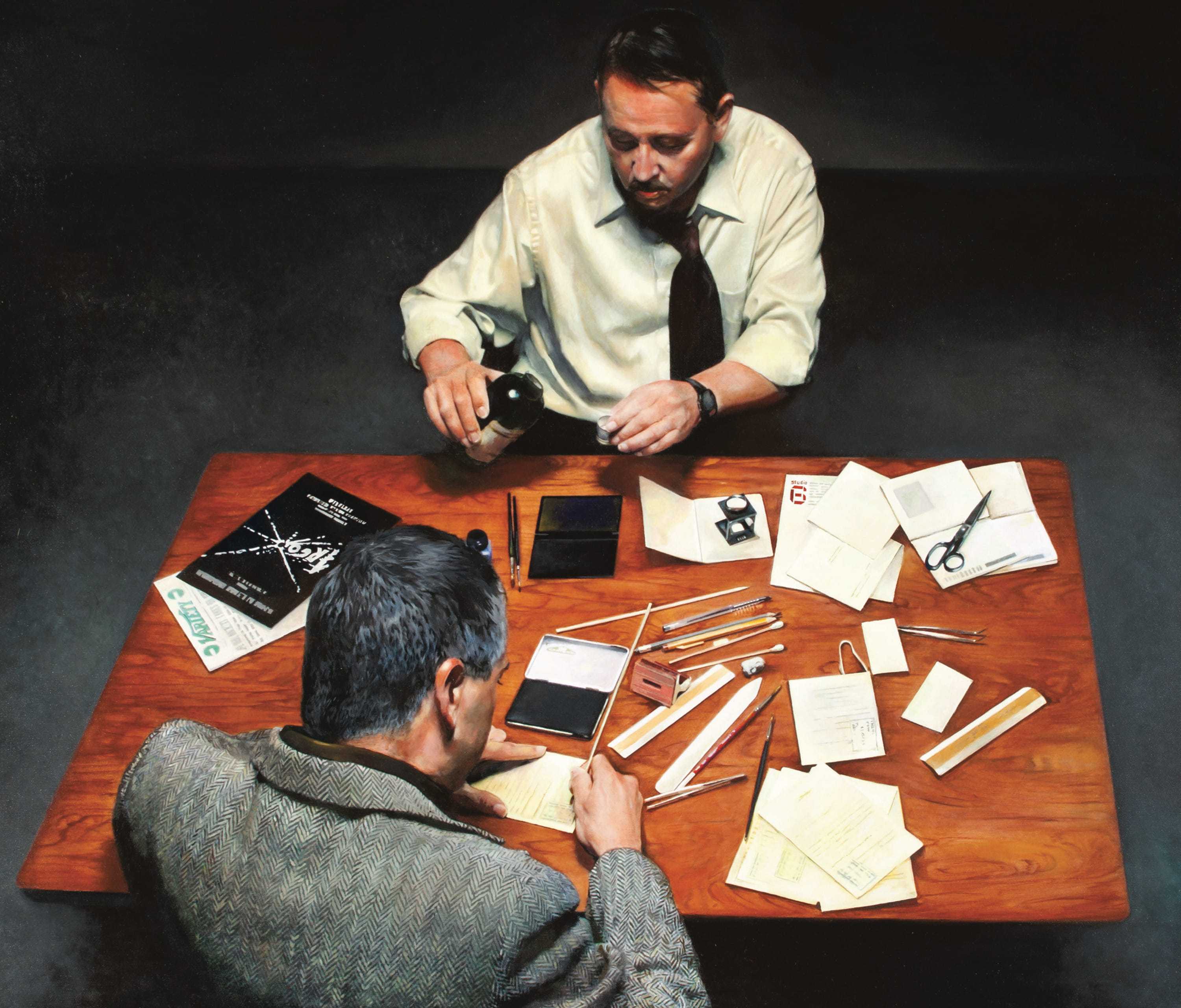 Two men sitting across from each other at a wood table scattered with papers and other objects.